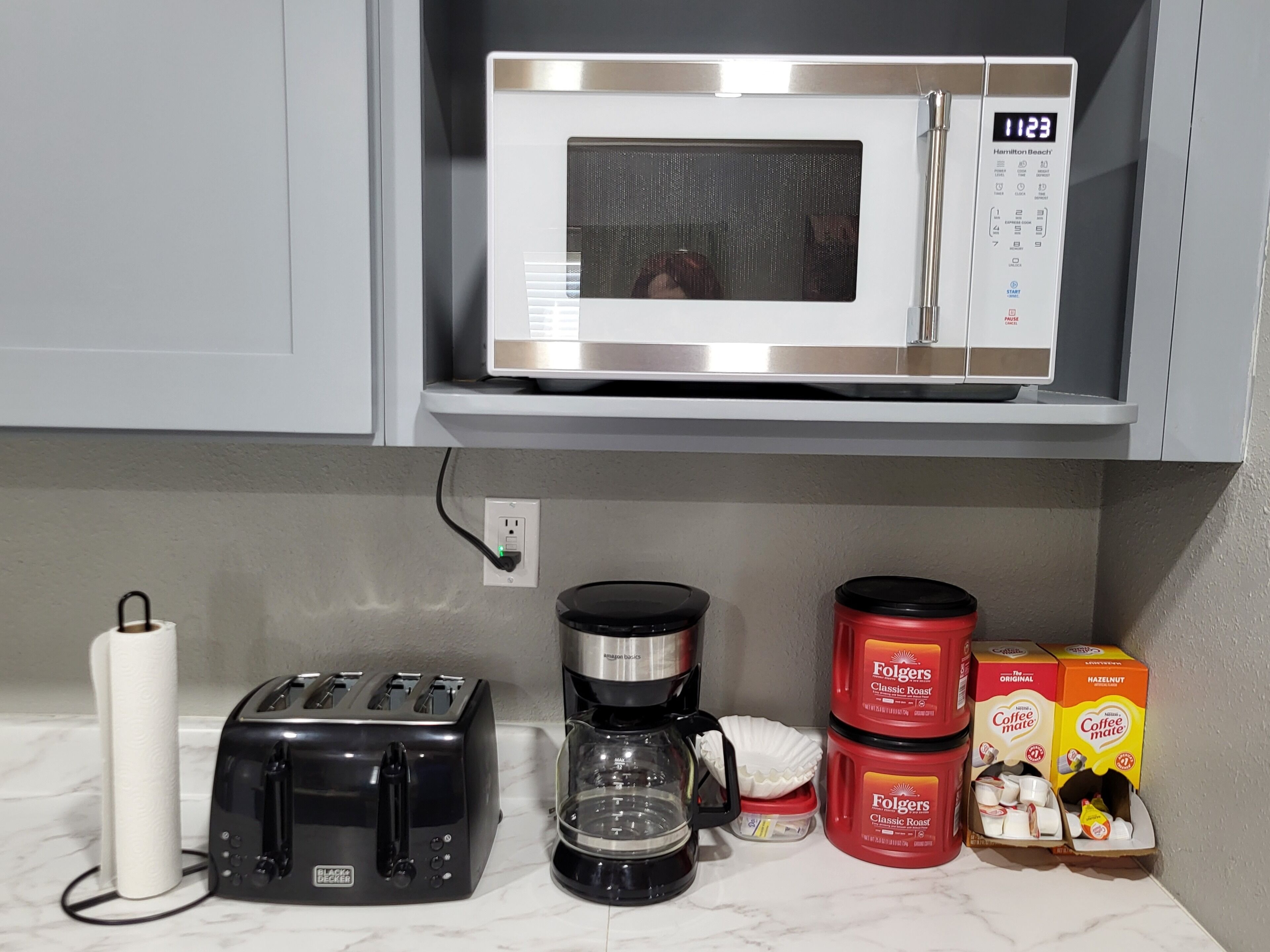 A kitchen with a microwave, toaster, coffee maker, and Folgers coffee boxes.
