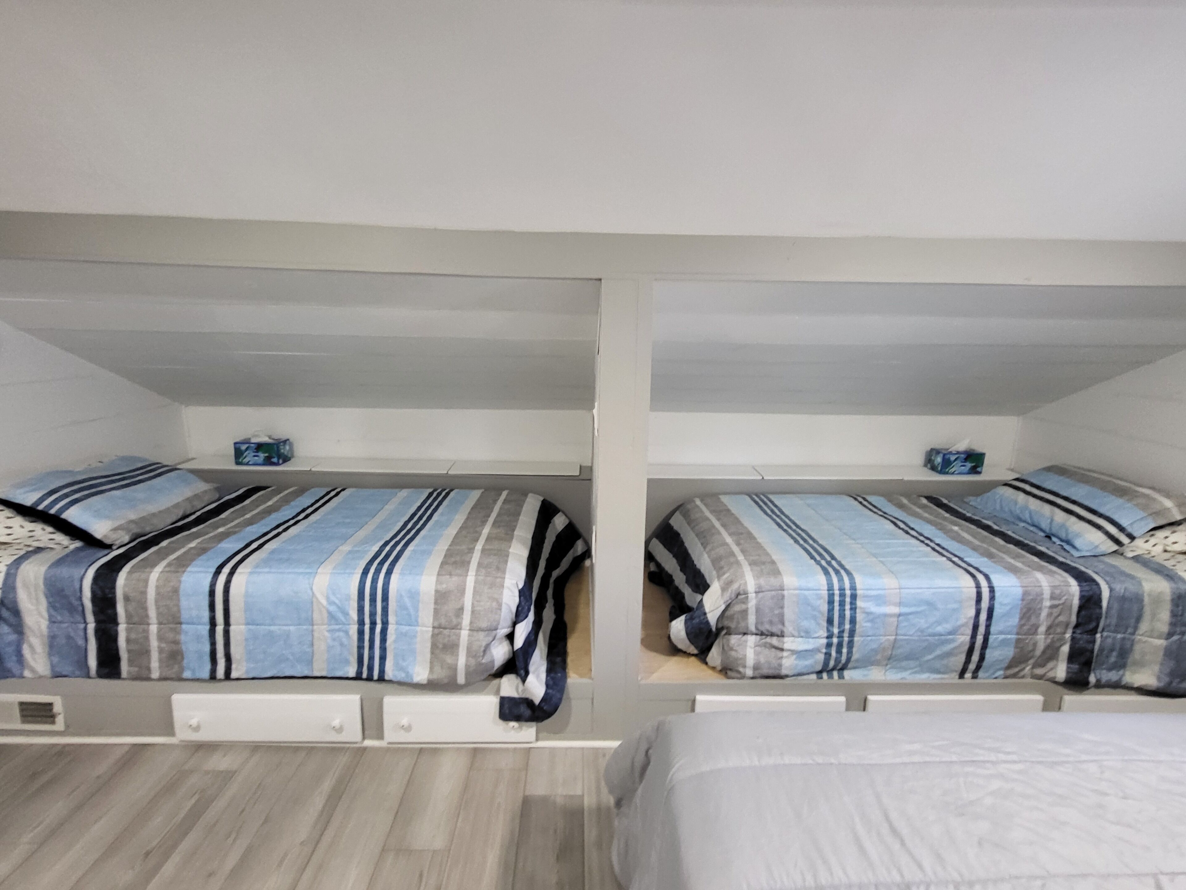 A room with two bunk beds, each with a striped bedspread and pillows. There are storage drawers under the beds and a small shelf with a box on the wall.