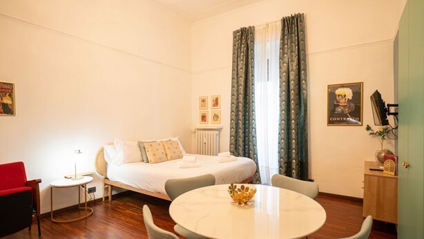 A hotel room with a bed, a round dining table, a chair, a small side table, and a red chair. - Crocetta Golden Soul | Design Loft near Porta Nuova (TORINO)