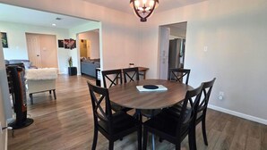 Uma sala de jantar com uma mesa redonda, seis cadeiras, um candelabro e uma cozinha ao fundo.