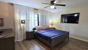 Um quarto com cama, mesas de cabeceira, ventilador de teto, televisão e janela com cortinas.