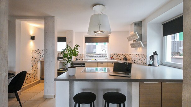 A modern kitchen with a central island, bar stools, a laptop, a potted plant, and a window with blinds.