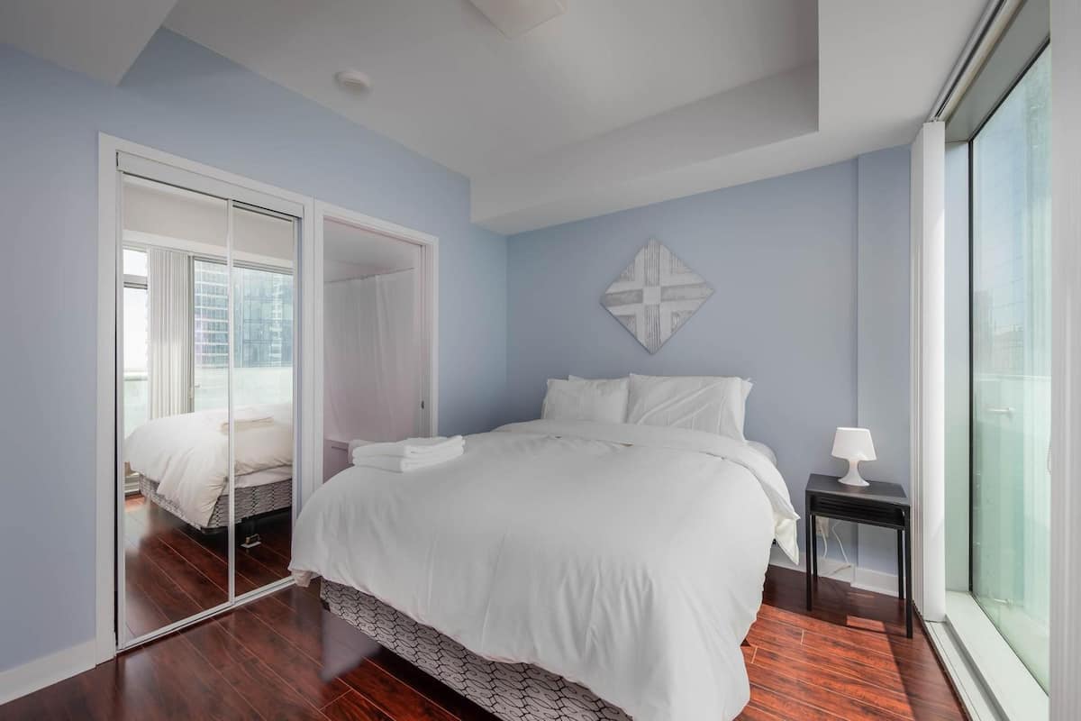 A bedroom with a large bed, a mirror, a bedside table with a lamp, and a view through a window.