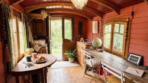 Une pièce chaleureuse avec un plafond en bois, une table à manger ornée d’un globe terrestre et d’un carnet, une fenêtre donnant sur un paysage verdoyant et un lustre.