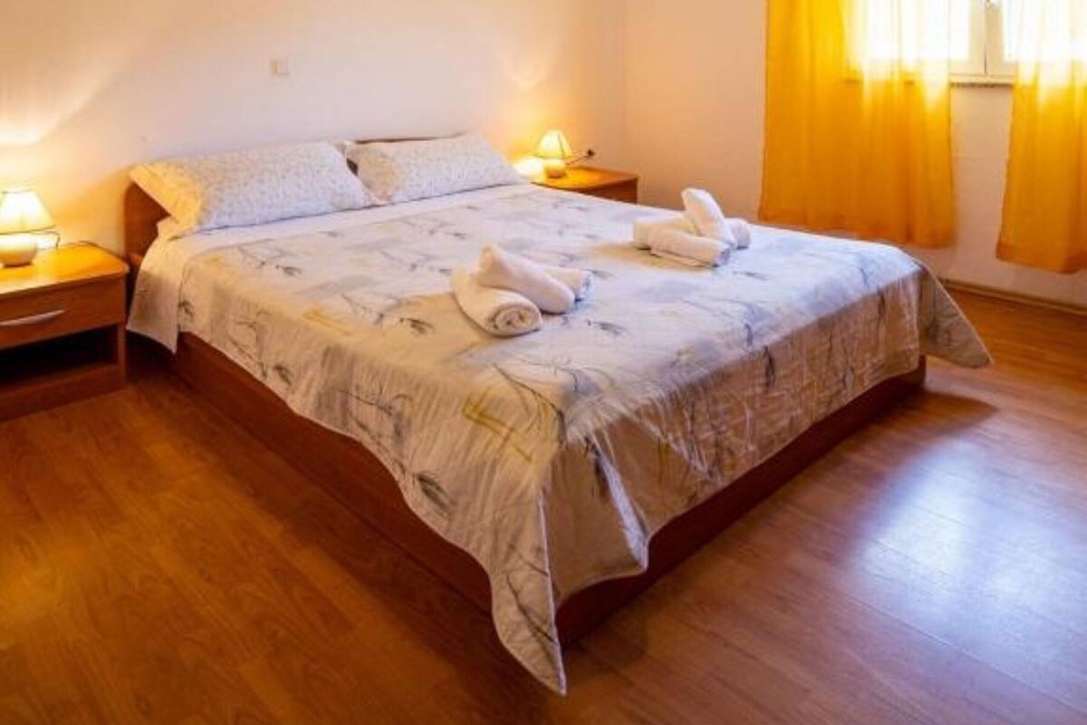 Ein Schlafzimmer mit Holzboden, einem Bett mit floralem Überwurf, zwei Nachttischlampen und einem Fenster mit gelben Vorhängen.