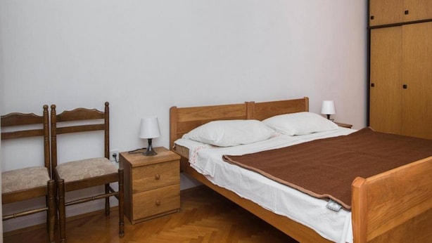 Uma cama de solteiro com cabeceira de madeira, mesa de cabeceira com luminária, cadeira de madeira e guarda-roupa.