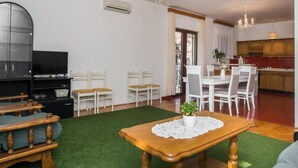 Sala de estar com televisão, canto de jantar com cadeiras, mesa de madeira e cozinha ao fundo.