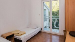Quarto pequeno e claro, com uma cama de solteiro, mesa de cabeceira de madeira, uma cadeira e uma porta de correr de vidro que dá para uma varanda externa.