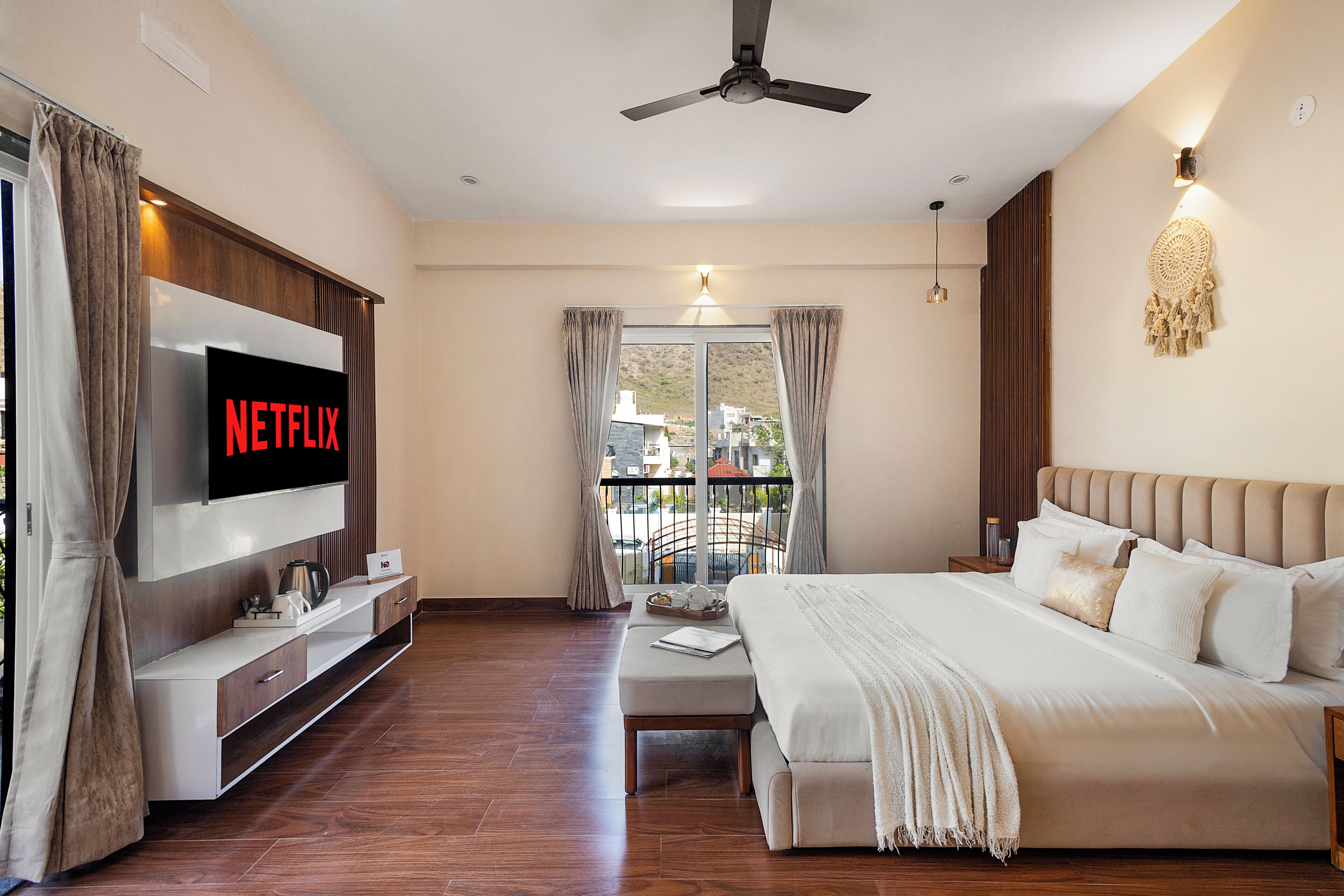 A hotel room with a bed, a TV displaying 'NETFLIX,' a ceiling fan, a balcony with curtains, and a bedside table.