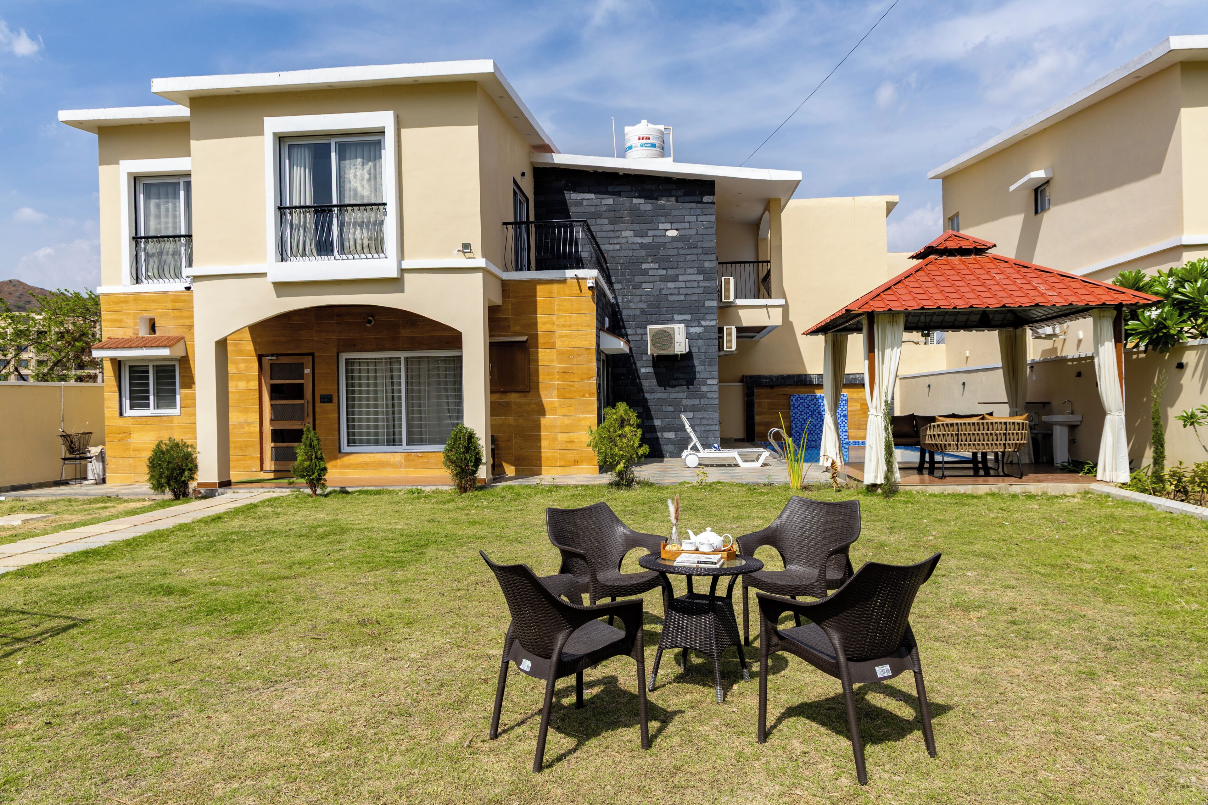 A modern two-story house with a flat roof, a balcony, and a small garden area with outdoor furniture.