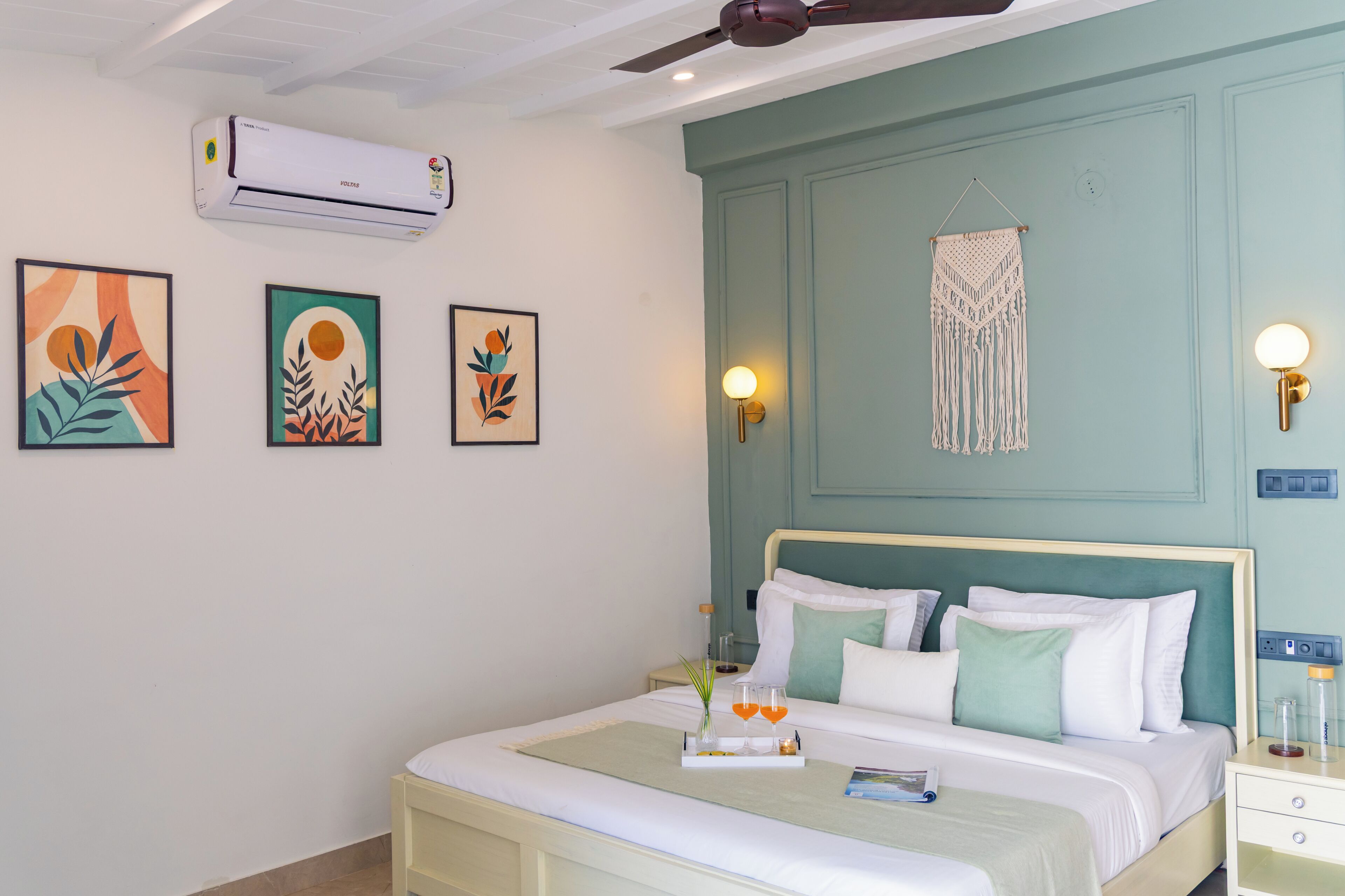 A bedroom with a bed, bedside tables, wall-mounted air conditioning, framed artwork, a macrame wall hanging, and a ceiling fan.