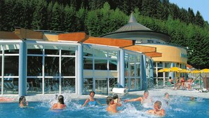 Un área de piscina con un edificio de cristal, una zona de estar cubierta y un fondo de bosque.