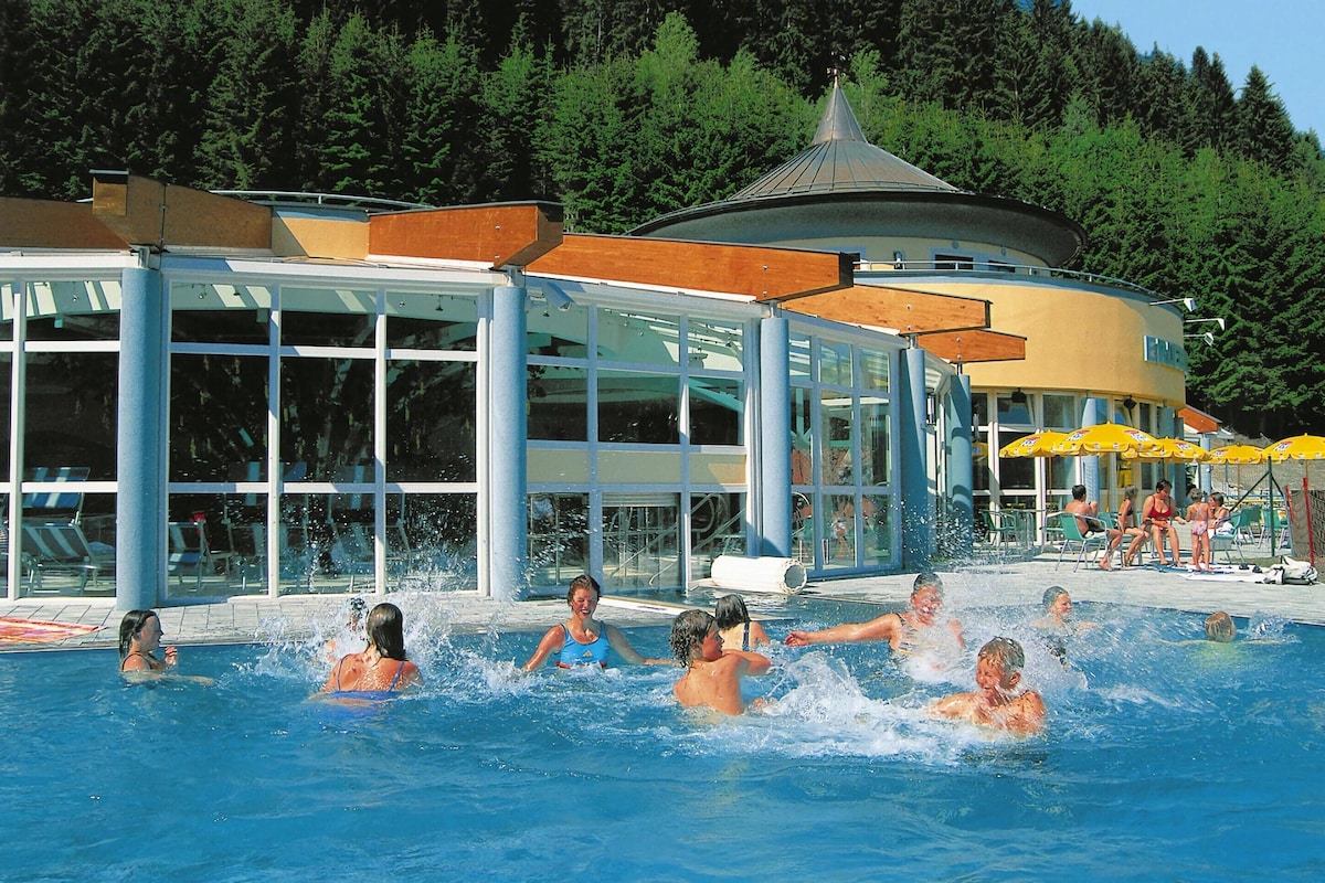 A swimming pool area with a glass-enclosed building, outdoor seating, and a forested backdrop.
