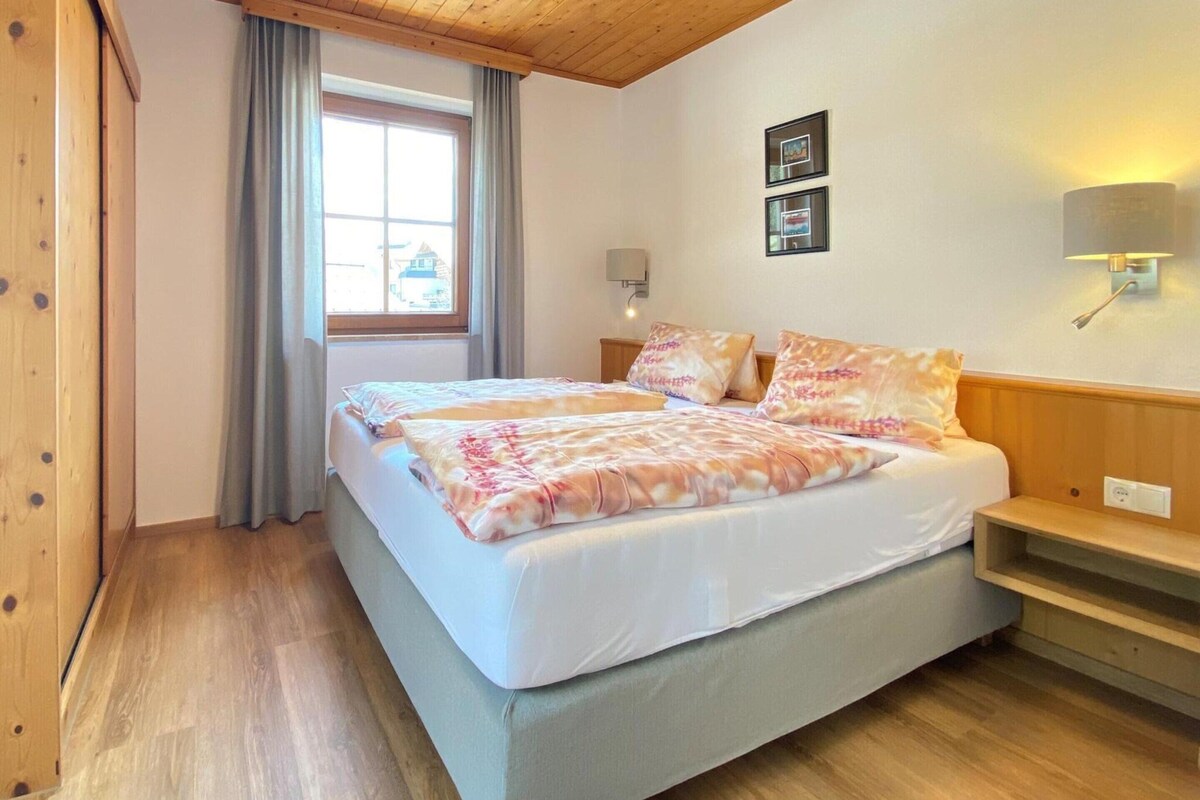 A double bed with a floral bedspread, a bedside table, a lamp, and a window with curtains.