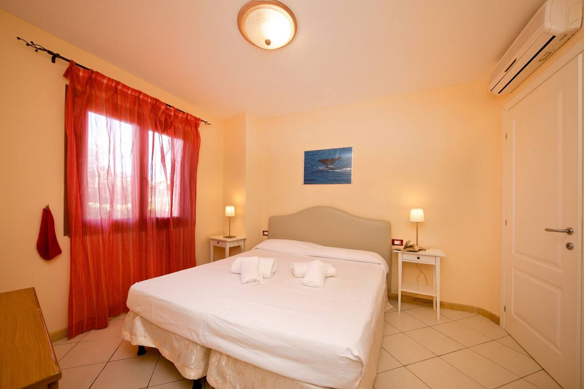 A hotel room with a bed, two bedside tables, a nightstand, a red curtain, a wall-mounted picture, and an air conditioning unit.