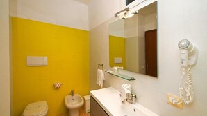 Casa de banho com parede amarela, lavatório branco, bidé, sanita e espelho. Secador de cabelo fixado na parede.
