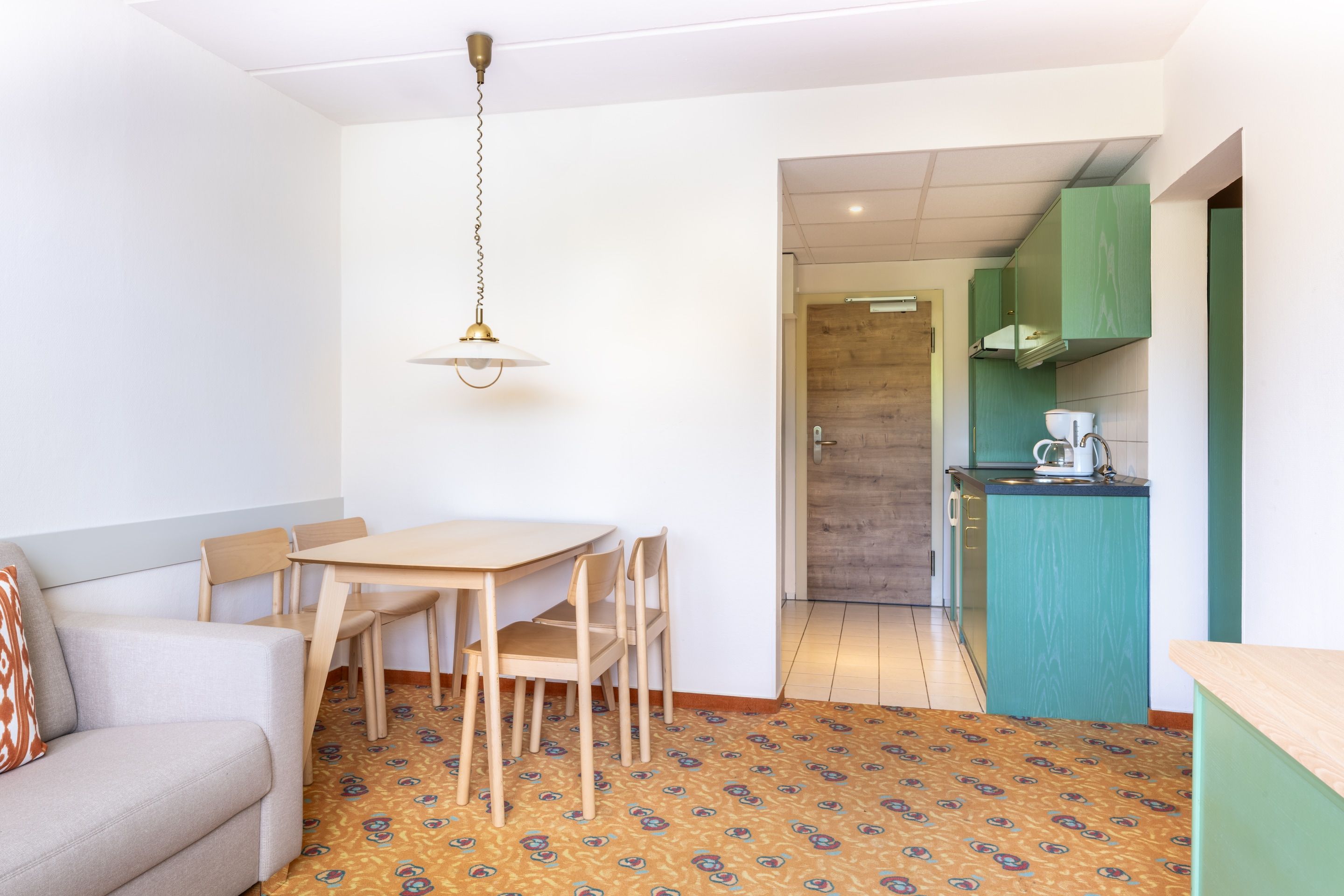 Ein modernes Zimmer mit Esstisch, Stühlen, einer Couch, einem gemusterten Teppich, einer Küchenzeile mit grünen Schränken und einer Pendelleuchte.