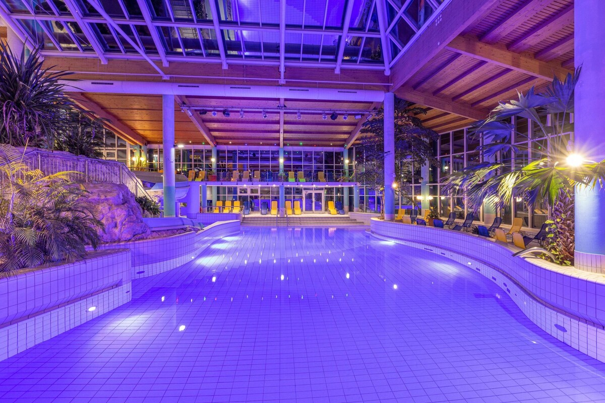An indoor swimming pool with a tiled floor, surrounded by plants and seating areas.