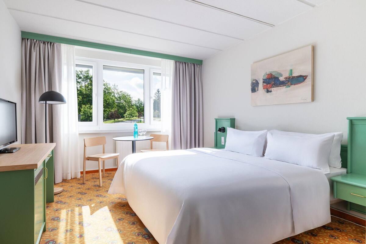 Une chambre d’hôtel avec un grand lit, un bureau, une chaise, une petite table et une fenêtre avec des rideaux.