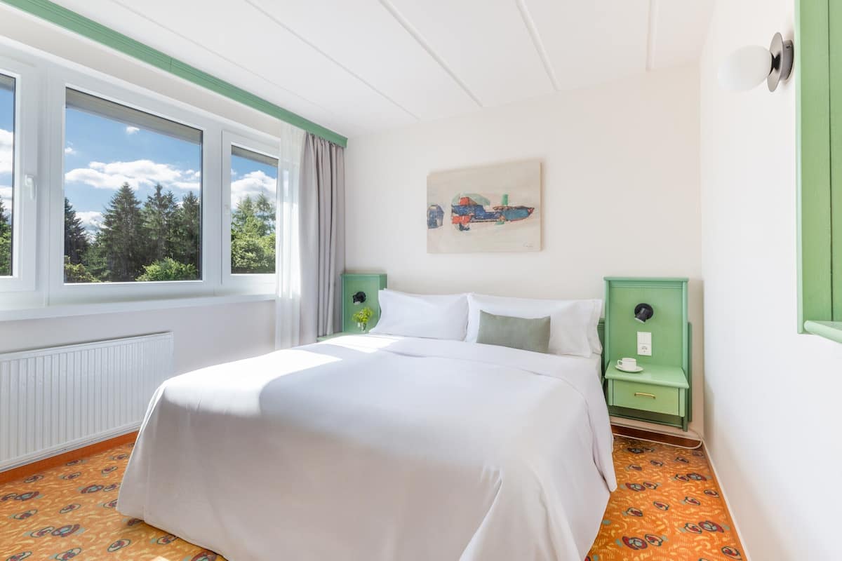 Une chambre d’hôtel avec un grand lit, une fenêtre avec des rideaux, une table de chevet verte et un tableau au mur.
