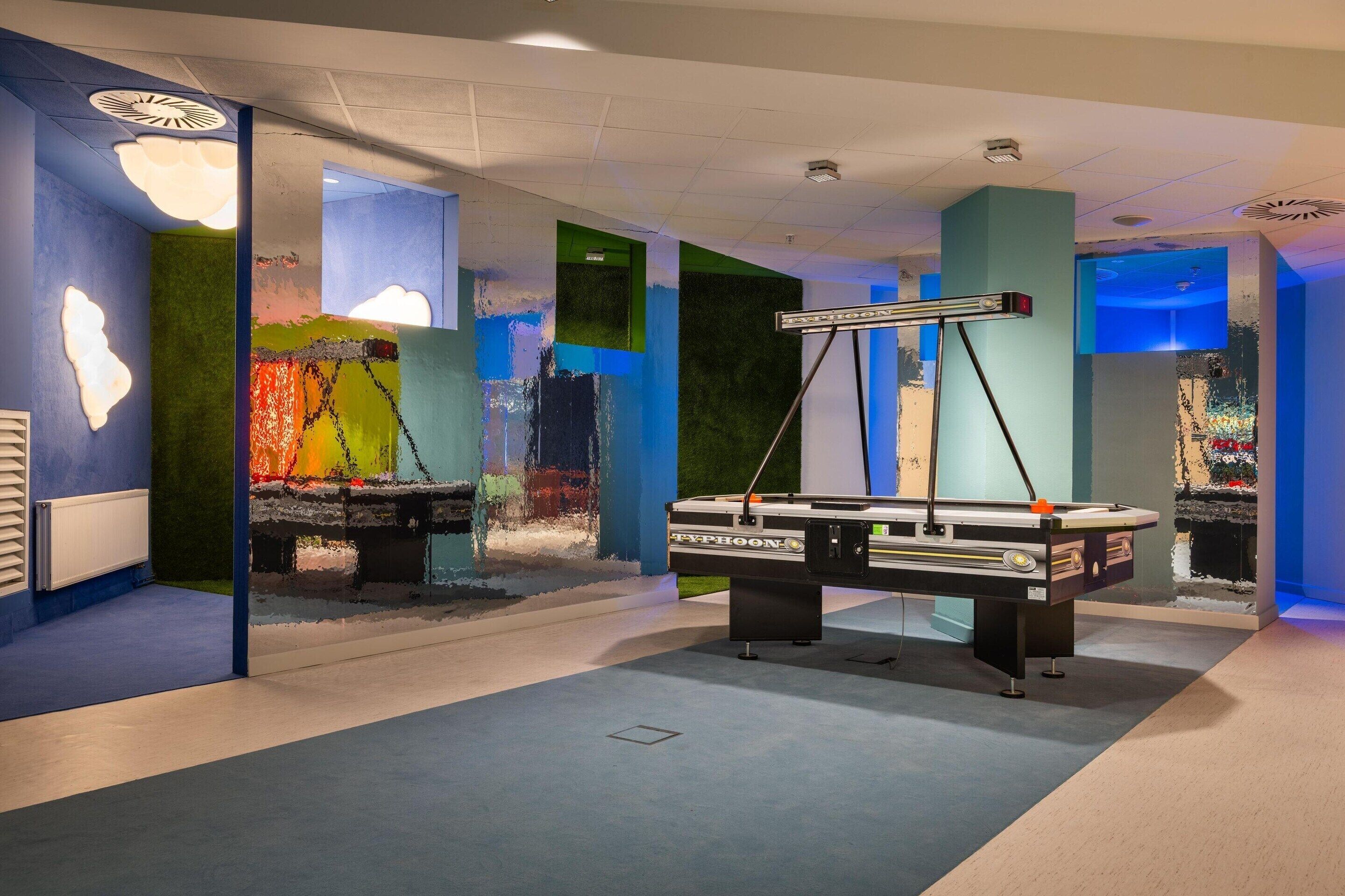 A room with a modern design, featuring a large table with a machine on it, possibly for printing or crafting. The walls are adorned with colorful abstract art.