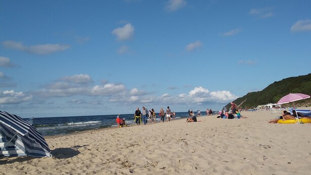 Una spiaggia con persone che camminano e siedono, un ombrellone a righe e una tenda sulla sabbia.