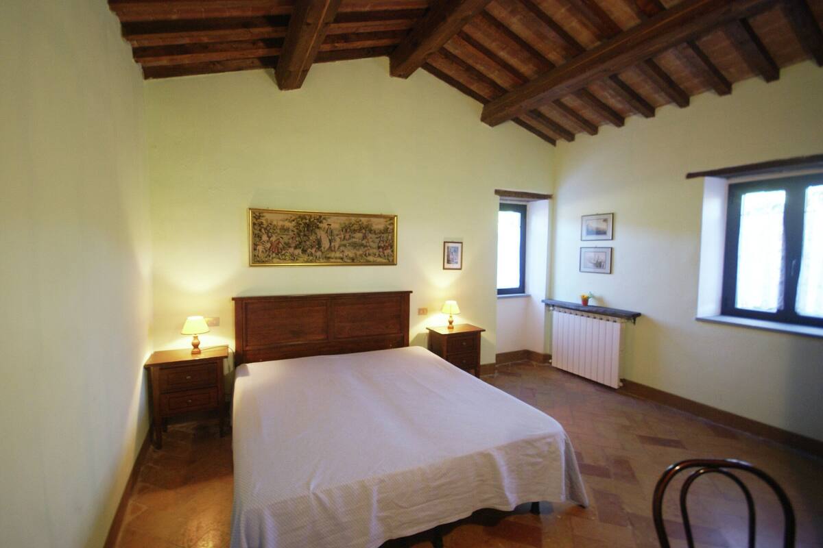 A bedroom with a wooden ceiling, a bed with a white bedspread, two bedside tables with lamps, a radiator, and a window with a view.