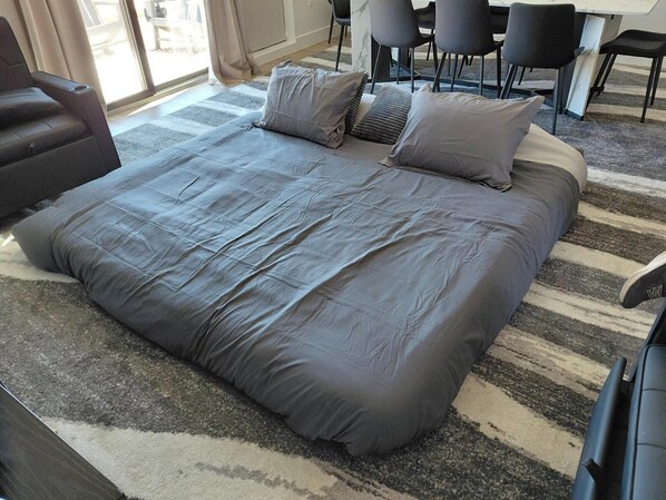 Un lit avec une literie grise, un tapis à rayures et un coin repas avec des chaises.