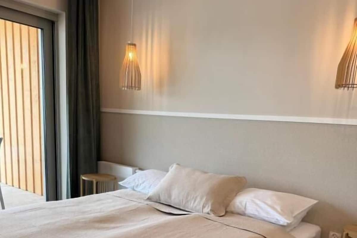 Ein ordentlich bezogenes Bett mit weißen Leinen, Holzboden und ein Fenster mit Vorhängen.
