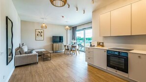 A modern living room with a sofa, a dining table with chairs, a TV, and a kitchen area with cabinets and an oven. - Stranda Residence Apartments Giżycko (Giżycko)