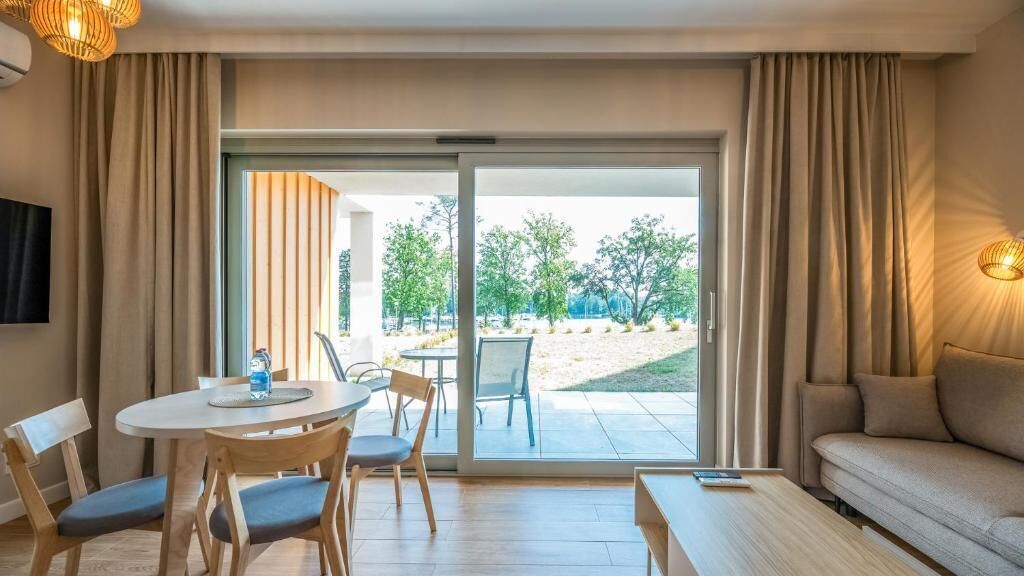 A hotel room with a dining area, a sofa, and a sliding glass door leading to an outdoor area with trees.