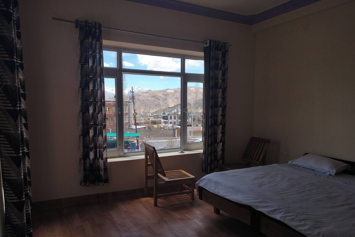 A bedroom with a bed, a chair, a window with curtains, and a view of mountains.