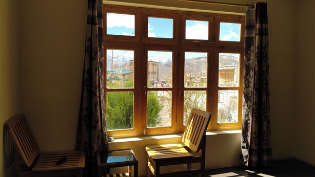 A room with a large window, a wooden chair, a small table, and a remote control on the chair. - Donskit guest house  (Leh)