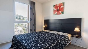 Ein Schlafzimmer mit einem Bett, zwei Nachttischlampen, einem Fenster mit Blick und einem Blumenbild an der Wand.