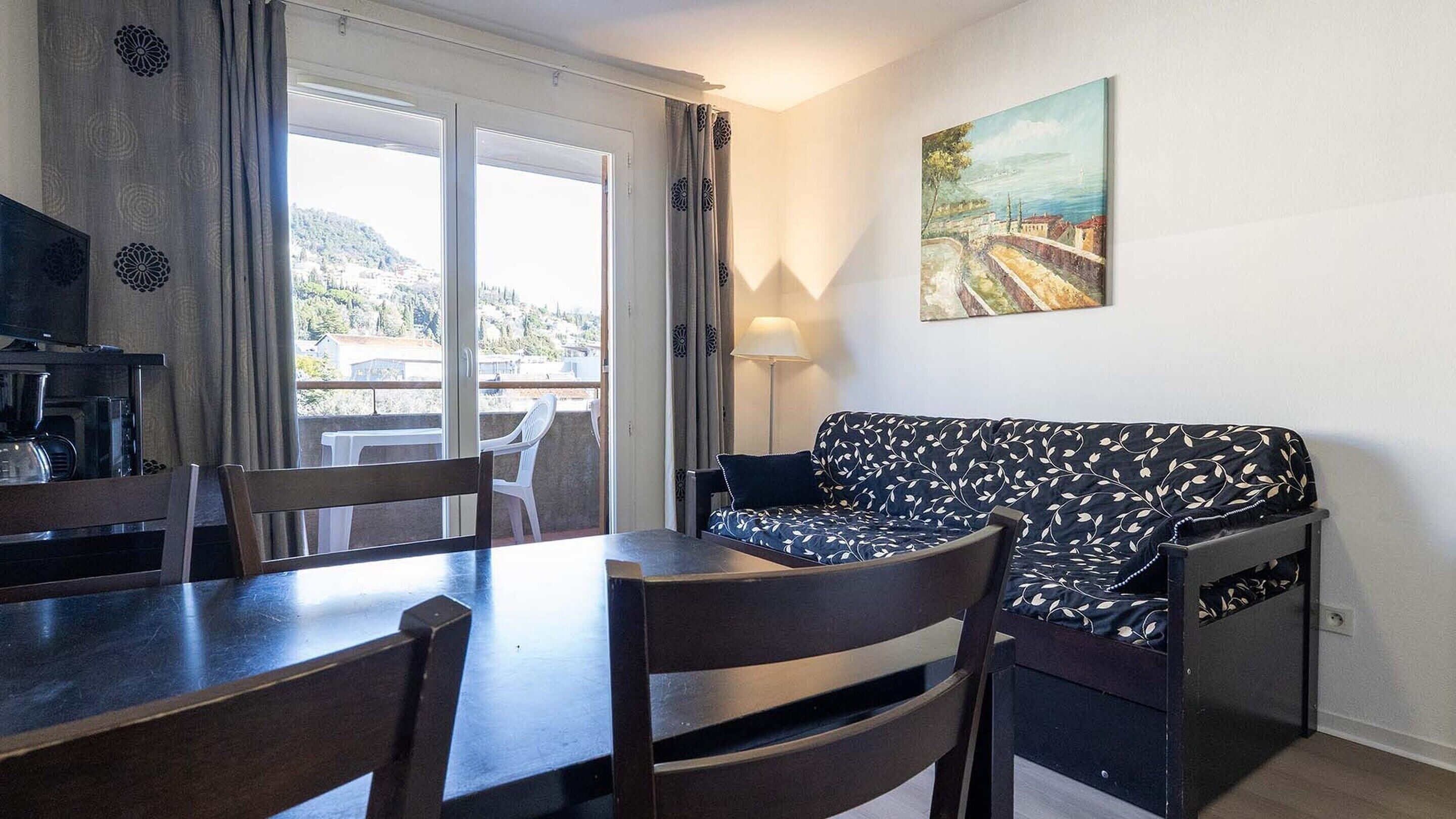 A room with a dining table, chairs, a sofa, a painting, and a view through a window.