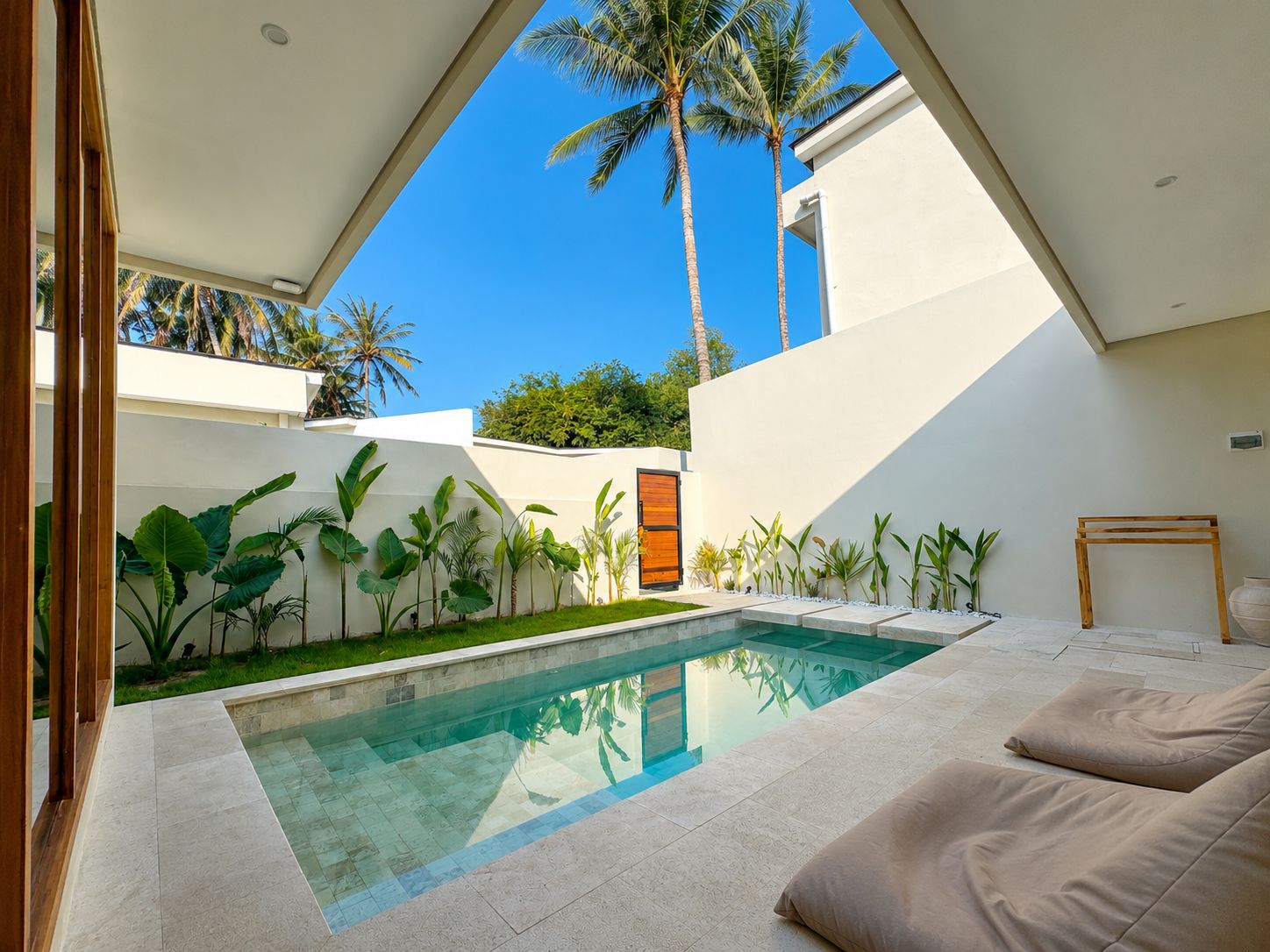 Uma área de piscina moderna, com piscina transparente, cercada por plantas tropicais e uma parede branca.