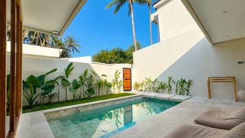 Uma área de piscina moderna, com piscina transparente, cercada por plantas tropicais e uma parede branca.