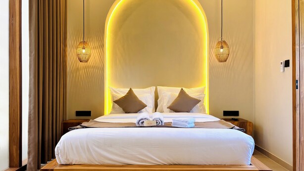 A hotel room with a large bed, two bedside tables, a wall-mounted control panel, and a wooden floor.