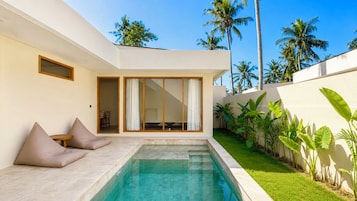 Un área de piscina moderna con una piscina cristalina, un espacio de descanso con cojines y vistas a un paisaje tropical con palmeras.