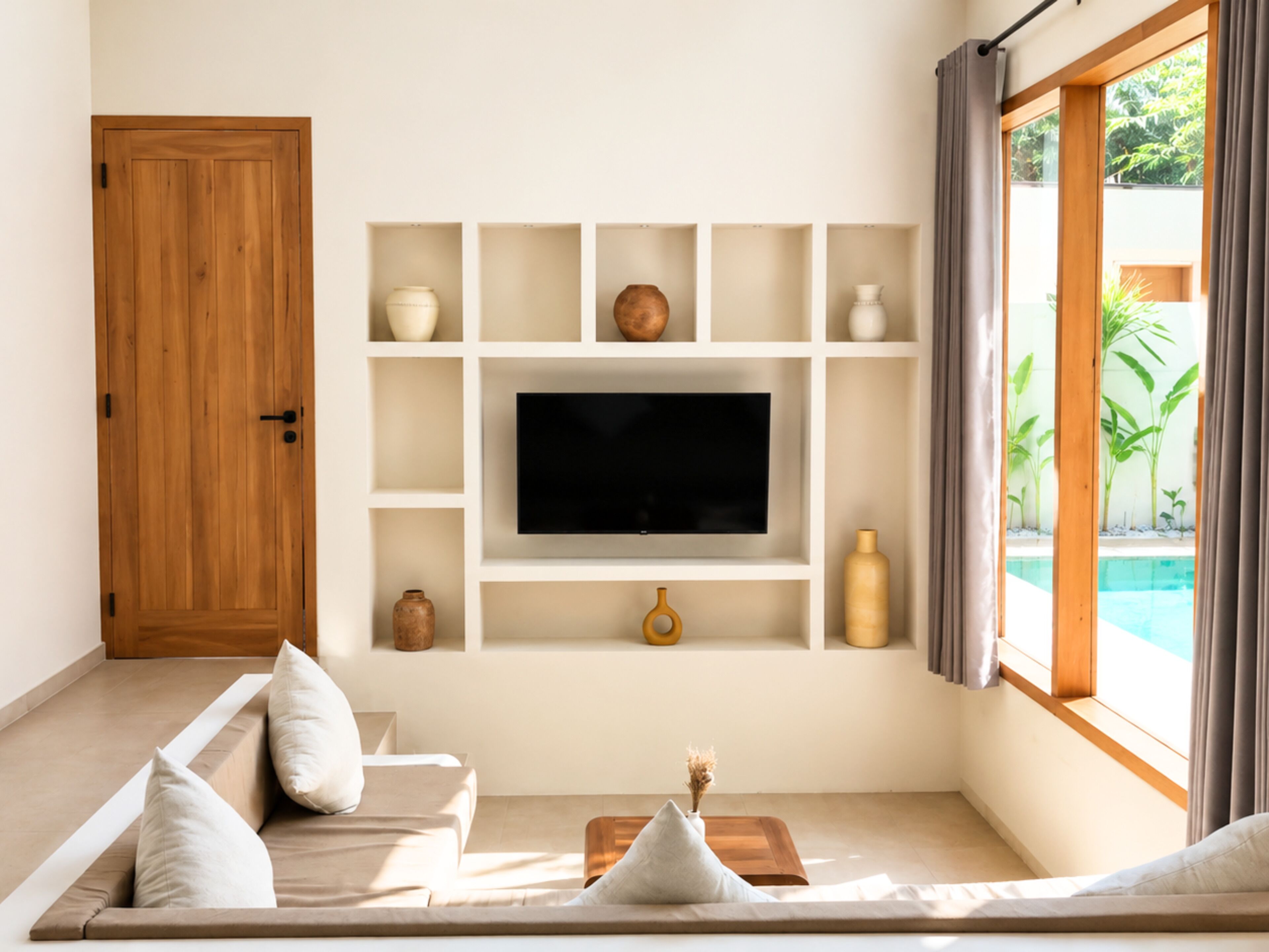 A modern living room with a flat-screen TV, built-in shelves, a wooden door, and a view of a swimming pool.