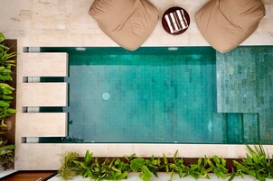 Una piscina rectangular con borde de azulejos, rodeada de plantas y cojines color beige.