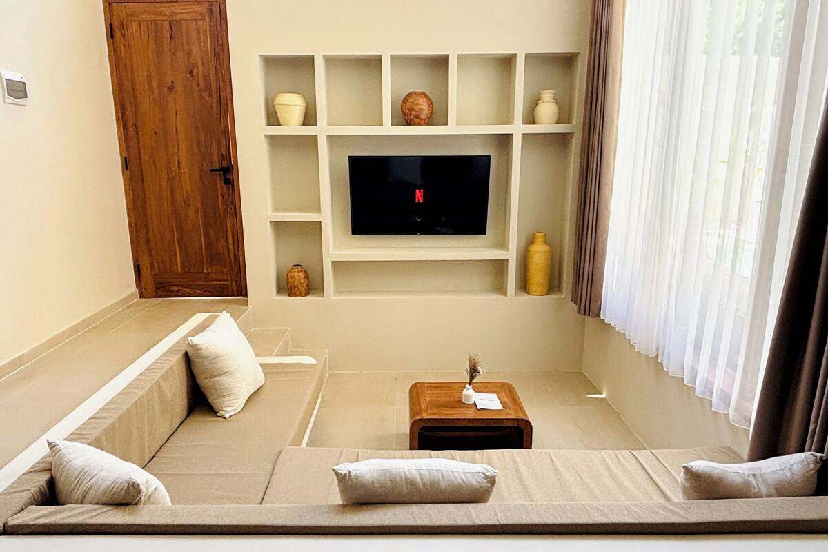 A living room with a sofa, a TV mounted on a wall, a wooden door, and a window with curtains.