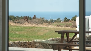 A view through a window of a grassy area with a stone wall, a wooden picnic table, and a white chair.