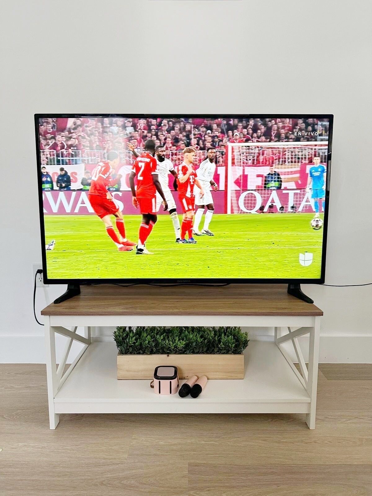 Un téléviseur diffusant un match de football, posé sur un support blanc, avec une plante et un petit tiroir en dessous.