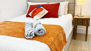 Un letto rifatto con cura, coperto da un piumone bianco e una coperta arancione, con asciugamani arrotolati. Un comodino in legno con una lampada è posizionato accanto al letto.