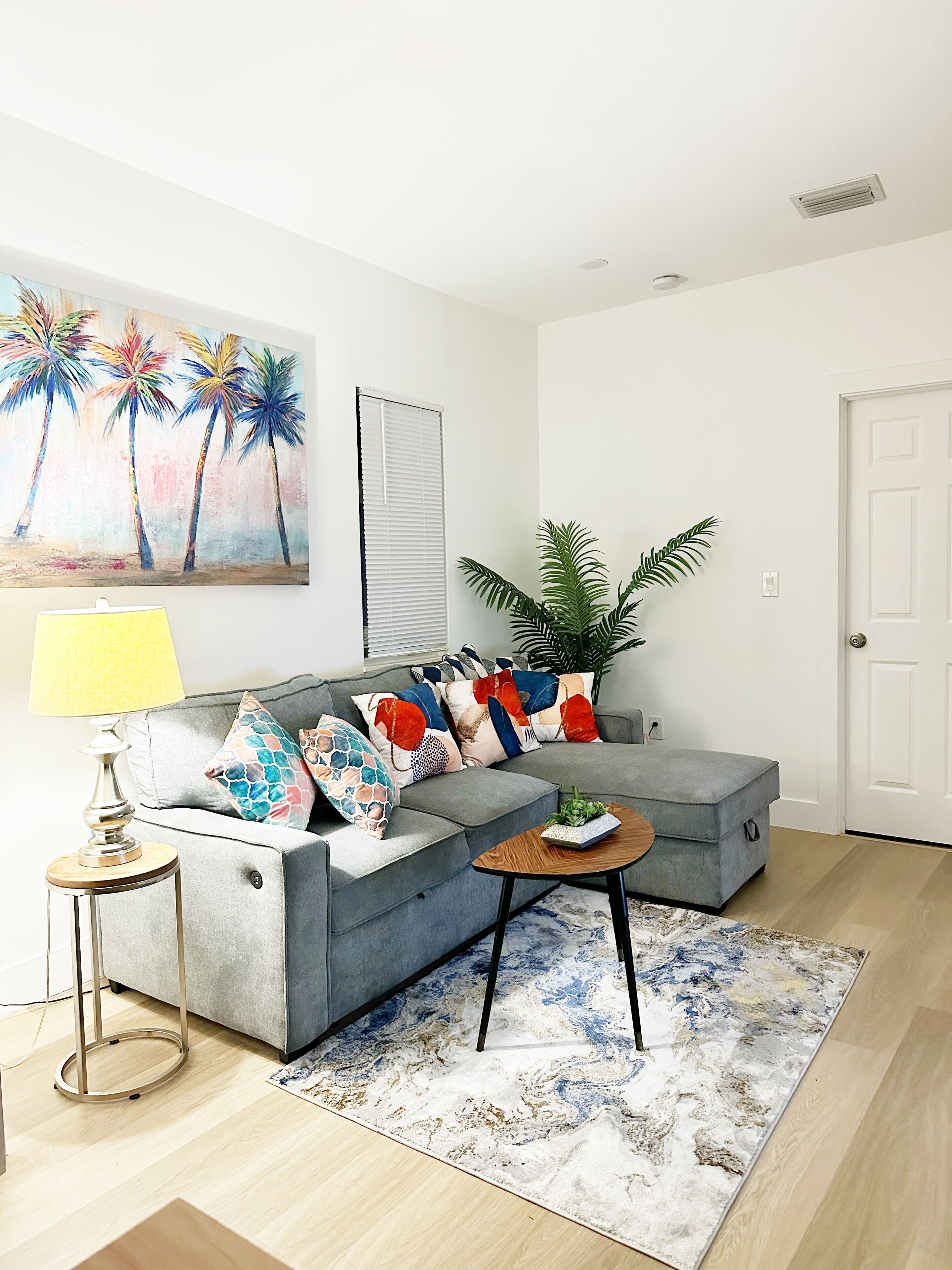Un salón con un sofá gris, almohadones coloridos, una mesita con una planta, una lámpara y un cuadro de palmeras en la pared.