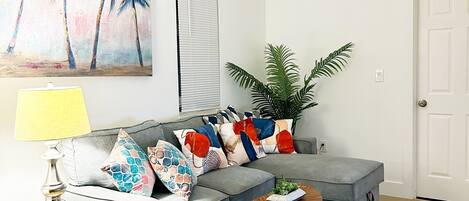 Un salón con un sofá gris, almohadones coloridos, una mesita con una planta, una lámpara y un cuadro de palmeras en la pared.