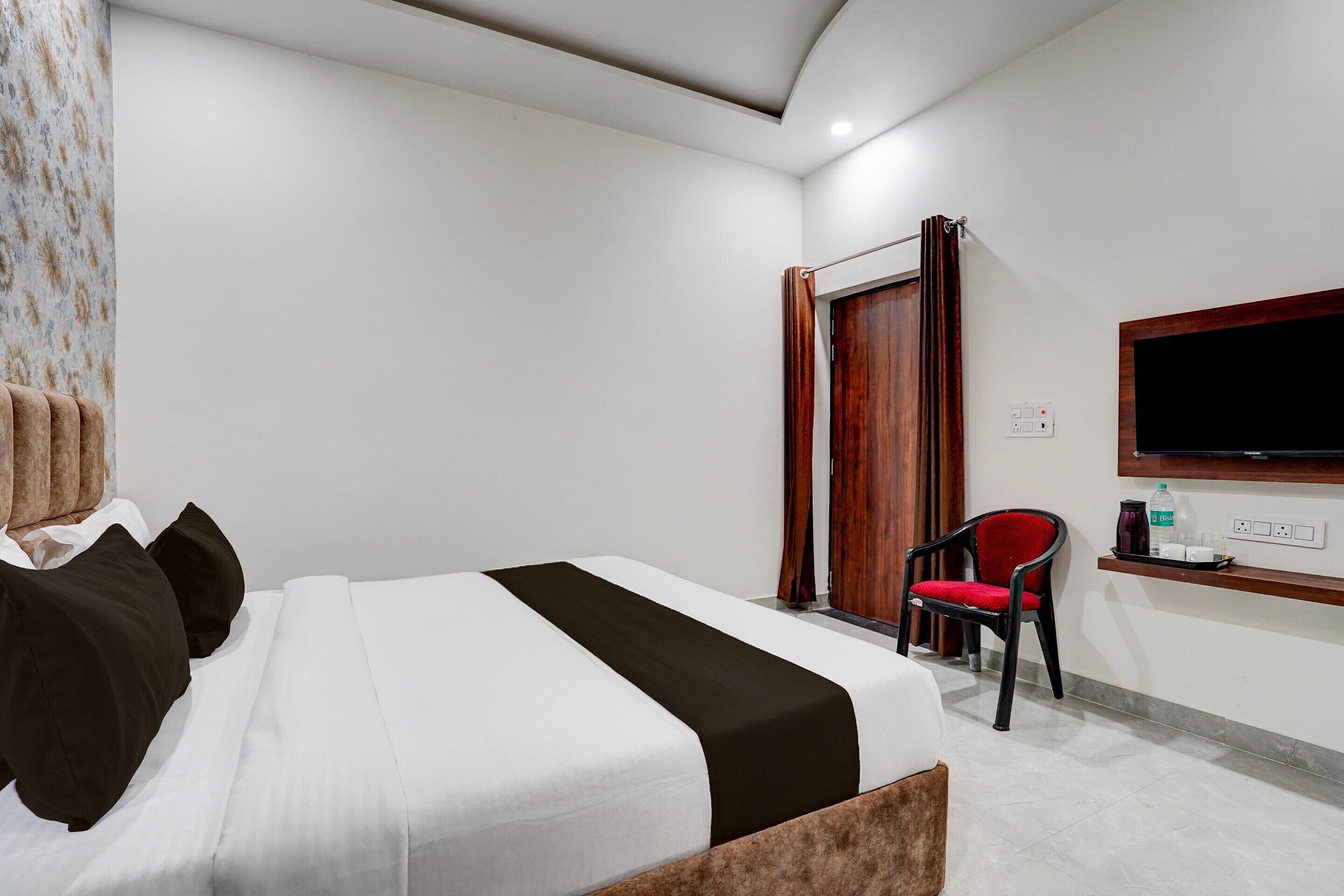 A hotel room with a bed, a TV mounted on the wall, a chair, and a small shelf with items.