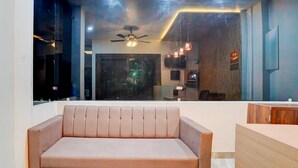 A modern living area with a sofa, a ceiling fan, pendant lights, and a view through a large window. - Premium Neelam Hotel (Bareilly)