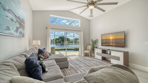 Ein Wohnzimmer mit Sofa, Fernseher, Deckenventilator und Seeblick durch Glastüren.